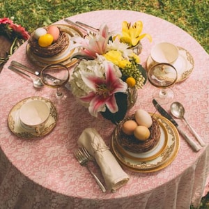May include: A pink tablecloth with a floral pattern is set for a spring-themed dinner party.  The table is decorated with a bouquet of white and yellow lilies, a nest of colorful eggs, and gold-rimmed plates and cups.