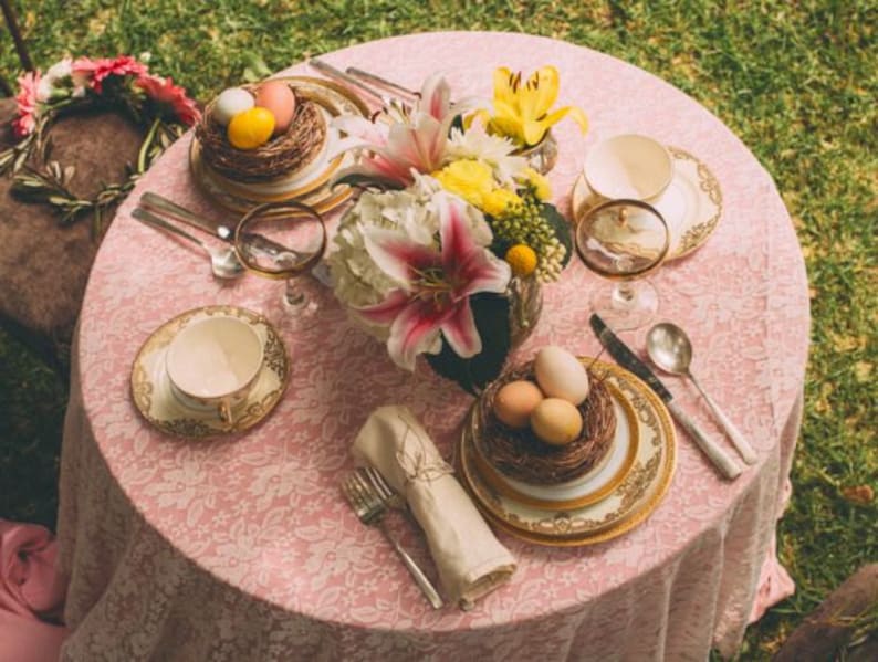 May include: A pink tablecloth with a floral pattern is set for a spring-themed dinner party.  The table is decorated with a bouquet of white and yellow lilies, a nest of colorful eggs, and gold-rimmed plates and cups.