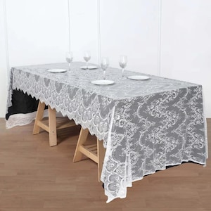 May include: White lace tablecloth with a delicate floral pattern. The tablecloth is draped over a table with four glasses and plates.