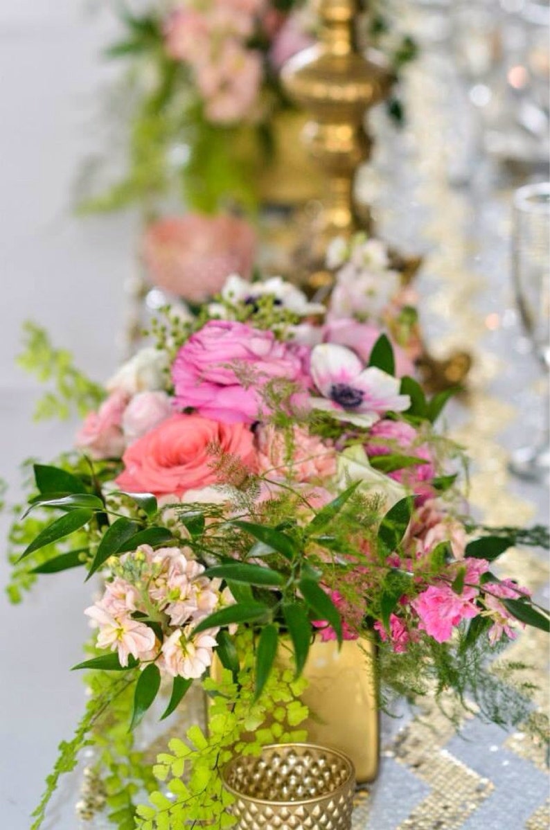 Puede incluir: Un arreglo floral en un jarr&oacute;n dorado con flores rosas, blancas y verdes. Las flores est&aacute;n dispuestas en un estilo moderno con vegetaci&oacute;n y helechos.