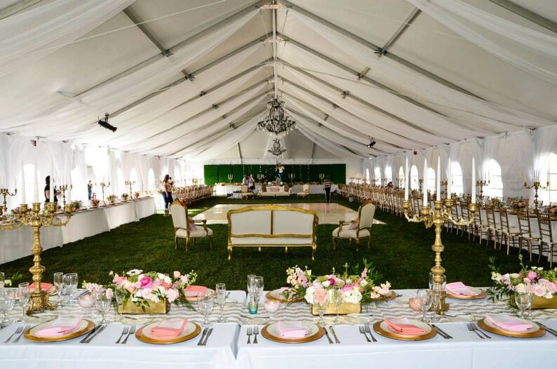 Puede incluir: Una carpa blanca con una pista de baile en el centro, decorada con manteles blancos, candelabros dorados y centros de mesa florales rosas. Las mesas est&aacute;n dispuestas con cargadores dorados, platos blancos y servilletas rosas.