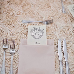 May include: A place setting with a white napkin, two forks, two knives, and a menu card with the letter "G" and the word "Menu" printed on it. The table is covered in a beige fabric with a rose pattern.