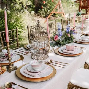 May include: A table setting for a party or special occasion with gold chargers, pink and white floral china, and pink candles. The table is set with a white tablecloth and gold flatware. There are flowers in a vase in the center of the table.