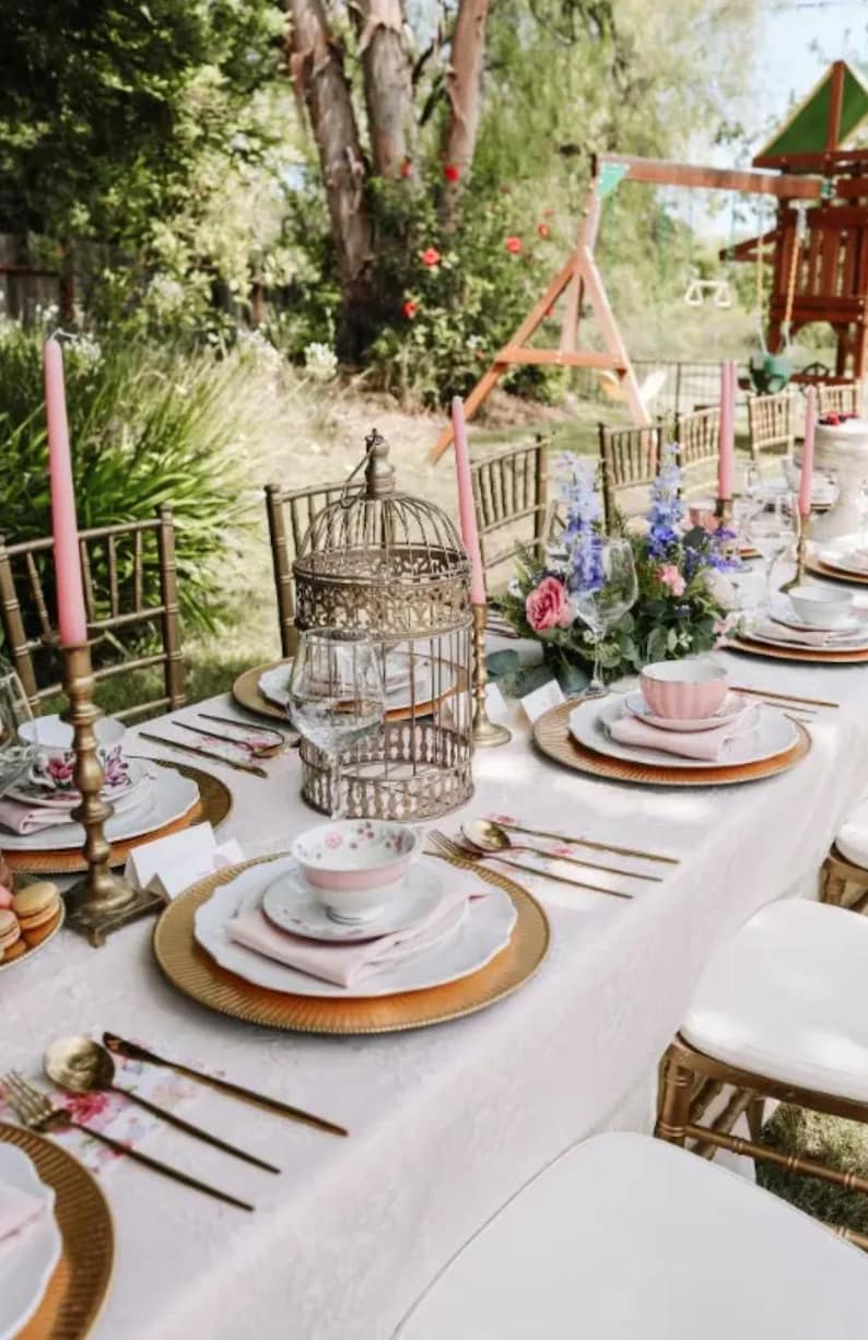 May include: A table setting for a party or special occasion with gold chargers, pink and white floral china, and pink candles. The table is set with a white tablecloth and gold flatware. There are flowers in a vase in the center of the table.