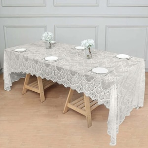 May include: White lace tablecloth with scalloped edges, laid on a wooden table with two wooden sawhorses. The tablecloth is decorated with a delicate floral pattern and has a subtle sheen.