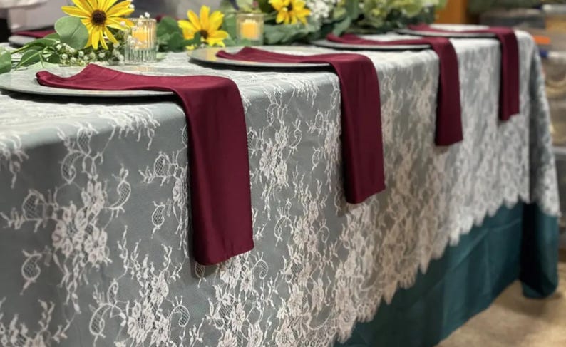 May include: A gray lace tablecloth with a teal border. Four burgundy napkins are placed on the table, with a white plate and a clear glass in front of each napkin.