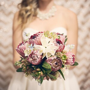May include: A bride holds a bouquet of pink and white flowers. The bouquet includes proteas, orchids, and greenery.