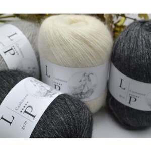 May include: Four balls of soft, fuzzy yarn in shades of cream, light gray, and dark charcoal gray. Each ball is wrapped in a white paper label featuring the text LP Cashmere, five stars, and a circular illustration of a goat.
