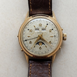 May include: A gold-tone Nivada Lamon automatic wristwatch with a textured off-white dial, featuring a moon phase indicator, day and month windows, and a date pointer hand. The watch has a dark brown leather strap with a floral embossed pattern.
