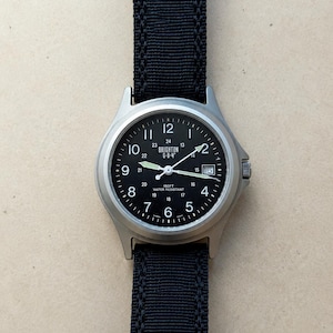 May include: A wristwatch with a round silver-tone metal case, a black dial featuring white Arabic numerals, a date window at the three o'clock position, and luminous hands. The watch includes a black nylon strap and displays the text BRIGHTON G-B-4.