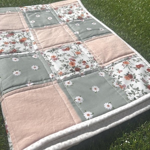 May include: A patchwork quilt featuring a repeating pattern of solid light pink squares, sage green squares with a white daisy print, and white squares with a multi-colored floral pattern. The quilt has a white border with a small grey cross pattern.