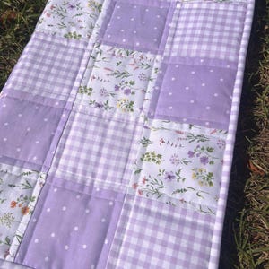 May include: A patchwork quilt featuring a repeating pattern of square fabric blocks. The design includes light purple fabric with white polka dots, a light purple and white gingham check, and a white fabric printed with small colorful wildflowers.
