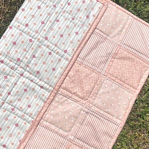 May include: A rectangular quilted runner featuring two distinct sections. One side displays a white fabric with thin vertical stripes and small pink and orange flower motifs. The other side consists of a patchwork of light pink floral and striped fabrics.