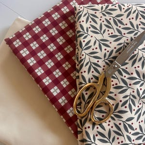 May include: Three folded pieces of fabric including one solid cream textile, one red and white gingham plaid textile, and one cream textile with a dark green leaf and red berry pattern, accompanied by gold-handled scissors labeled Ever SharpGold.