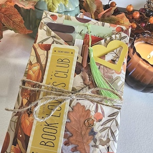 May include: A rectangular gift package wrapped in autumn-themed paper with pumpkins and leaves, tied with twine. It features a yellow bookmark labeled BOOK LOVERS CLUB, a light green tassel, a yellow heart-shaped cutout, and a dried oak leaf accent.