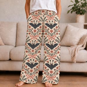 Op de afbeelding: Wijde loungebroek met een crèmekleurige achtergrond en een herhalend patroon van donkergrijze vleermuizen, roodbruine bloemen, groene bladeren en lichtrode strikken. De broek heeft een elastische tailleband en zijzakken, gecombineerd met een wit T-shirt.