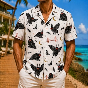 May include: A short-sleeved button-down shirt in off-white featuring an all-over print of black moths with red eyes, golden crescent moons, small stars, feathers, and red illustrations of the Golden Gate Bridge across the fabric.