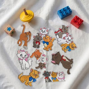 May include: A collection of embroidered patches featuring various cartoon cats, including orange, white, and grey kittens with colorful bows and collars, alongside a yellow spinning top, a blue building block, and a red building block.