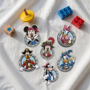 Op de afbeelding: Zes geborduurde patches met Disney-figuren verkleed als piraten, waaronder Mickey, Minnie, Donald, Daisy, Goofy en Pluto, elk ingelijst in een grijs patrijspoortontwerp, vergezeld door een gele tol en twee plastic bouwstenen.