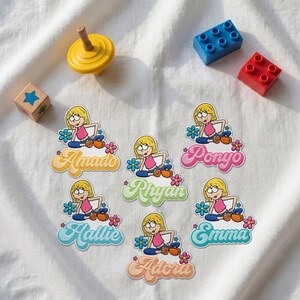 Könnte beinhalten: Sechs gestanzte Aufkleber mit einem Cartoon-Mädchen mit blonden Haaren, rosa Oberteil und blauer Hose, jeweils mit einem Namen in Schreibschrift: Amado, Ponyo, Rhyan, Hallie, Emma und Adora. Umgeben von einem gelben Kreisel und bunten Bausteinen.