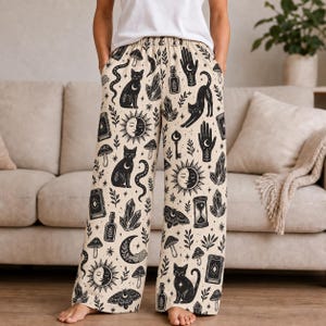 Op de afbeelding: Wijde loungebroek in een crèmekleur met een all-over zwarte grafische print van katten, zonnen, manen, kristallen, paddenstoelen, motten, handen, sleutels, zandlopers en tarotkaarten. De broek heeft een elastische tailleband en zijzakken.