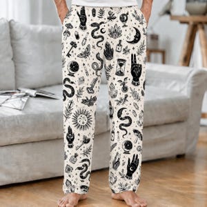 Op de afbeelding: Off-white loungebroek met een all-over zwarte inktstijl print van mystieke symbolen, waaronder slangen, kristallen, zonnen, manen, handen, zandlopers, sleutels, motten en paddenstoelen. De broek heeft een rechte snit en een elastische tailleband.