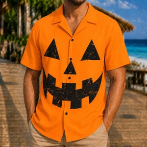 Puede incluir: Una camisa de color naranja brillante de manga corta con botones, que presenta un gráfico grande de una cara de calabaza de Halloween en negro impreso en la parte delantera. El diseño incluye dos ojos triangulares, una nariz y una boca.