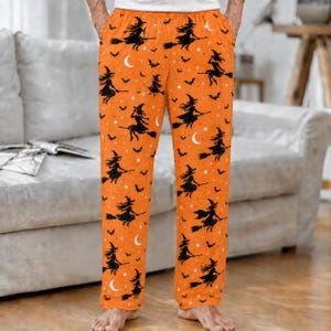 Op de afbeelding: Oranje pyjamabroek met een herhalend patroon van zwarte silhouetten van heksen op bezemstelen, kleine zwarte vleermuizen, witte maansikkels en witte sterren. De broek heeft een rechte pasvorm en een elastische tailleband.