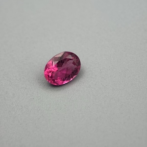 Puede incluir: Una piedra preciosa suelta de corte ovalado con un color rosa magenta intenso y una superficie facetada que refleja la luz. La piedra presenta una calidad transparente con inclusiones internas visibles y un acabado pulido.
