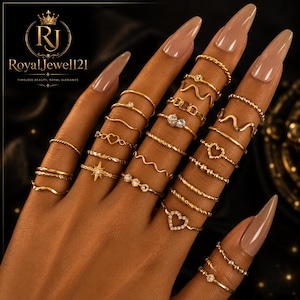 May include: A collection of gold-toned rings worn on fingers, featuring various designs including plain bands, twisted textures, heart shapes, wavy lines, small clear stones, and a starburst motif. The rings are arranged in multiple stacks.
