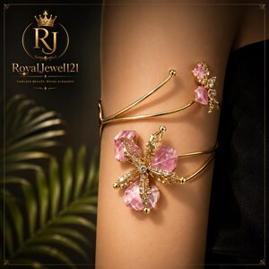 May include: A gold-toned arm cuff featuring a large floral motif adorned with faceted pink stones and small clear crystals. The design includes a secondary smaller floral accent at the top, with thin, flexible gold-toned metal bands wrapping around the arm.