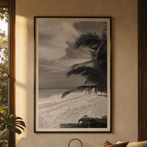 May include: A framed black and white wall art print depicting a tropical beach scene with palm tree fronds hanging over the sandy shore and ocean waves under a cloudy sky, presented in a thin dark frame with a wide white mat border.