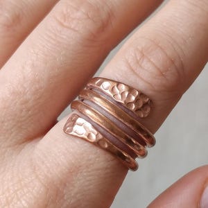 Peut inclure: Une bague en cuivre au design enveloppant à plusieurs anneaux. La bague se compose de trois bandes distinctes, les deux bandes extérieures présentant une finition martelée texturée, tandis que la bande centrale reste lisse et polie.