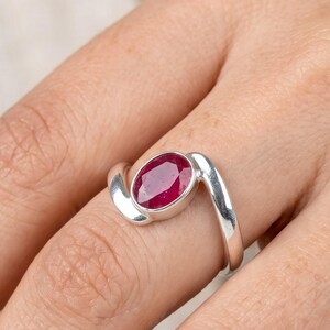 May include: A silver ring featuring a polished, oval-cut deep red ruby gemstone set in a smooth metal bezel. The band has a bypass design where the two ends of the silver shank curve around the central stone, creating a continuous, flowing look.