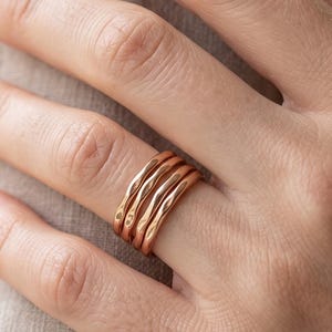 Peut inclure: Un ensemble de trois bagues empilées en or rose, chacune présentant une texture de surface lisse, ondulée et sinueuse. Les bagues sont fines et de largeur uniforme, créant un aspect superposé lorsqu'elles sont portées ensemble sur un doigt.
