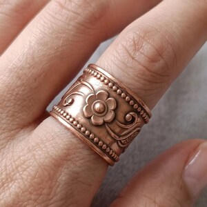 May include: A wide copper band ring featuring a raised floral motif in the centre, flanked by scrolling vine patterns and bordered by two rows of small, evenly spaced beaded textures along the top and bottom edges of the metal surface.