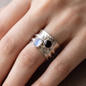 May include: A wide silver spinner ring featuring a textured, hammered finish. The band is adorned with two small, circular bezel-set gemstones: one iridescent, translucent moonstone and one solid, opaque black onyx stone side by side.