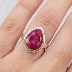 May include: A silver ring featuring a large, pear-shaped, faceted deep pink gemstone set in a smooth bezel. The metal band displays a series of small, vertical etched lines along the outer edge of the setting, creating a textured border around the stone.