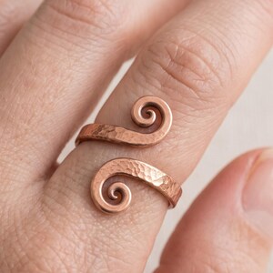 Peut inclure: Une bague en cuivre présentant un design ouvert à deux extrémités en spirale. La surface du métal présente une texture martelée distincte, créant une série de petites indentations sur l'anneau qui captent la lumière et offrent un fini tactile.