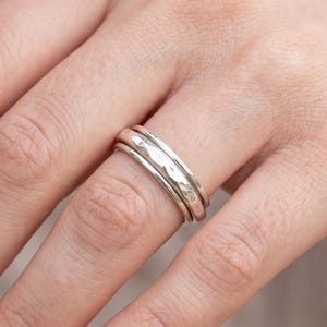 May include: A silver-coloured metal ring featuring a multi-band design. The central band has a textured, hammered surface, while the two outer bands are smooth and polished. The ring is worn on a finger, showcasing its layered, metallic construction.