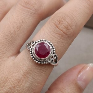 May include: A sterling silver ring featuring a round, faceted deep red gemstone set in a bezel. The band is decorated with a beaded texture and twisted rope detailing, with open heart-shaped accents on the sides of the central stone setting.