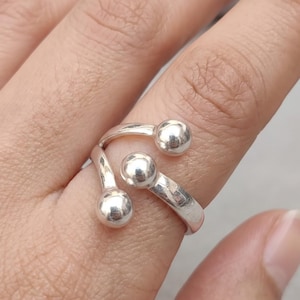 Könnte beinhalten: Ein silberfarbener Ring mit offenem Design und drei polierten, kugelförmigen Perlen an den Enden. Das Band schmiegt sich um den Finger und zeigt eine glatte, reflektierende Metalloberfläche an jedem der drei abgerundeten Kugelakzente.