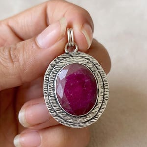 May include: An oval pendant featuring a faceted deep pink ruby gemstone set in a textured silver frame. The metal border has a dark, oxidised finish with horizontal etched lines surrounding the central stone and a small silver bail at the top.