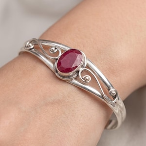 May include: A silver cuff bracelet featuring a central oval-shaped deep red ruby gemstone. The metal band displays a decorative openwork design with swirling wire accents and small spherical details on either side of the stone setting.