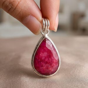 May include: A teardrop-shaped pendant featuring a faceted, deep magenta-coloured gemstone set in a polished silver frame. The pendant includes a silver bail at the top, designed for attachment to a necklace chain, showcasing a textured stone surface.