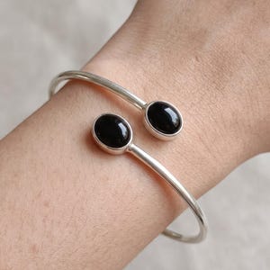 May include: A silver-toned open cuff bracelet featuring two polished, oval-shaped black stones at each end. The metal band is smooth and circular, designed to wrap around the wrist with the two black stones facing each other in the centre.