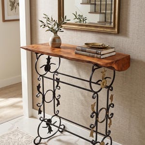 Op de afbeelding: Een consoletafel met een natuurlijk blad van hout met boomkant en een zwart metalen onderstel met decoratieve goudkleurige bloemaccenten. Op de tafel staan een kleine keramische vaas met groene takjes, twee boeken en een goudkleurig schaaltje.
