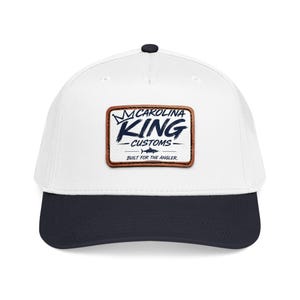 May include: A white baseball cap with a dark navy blue brim and a rectangular brown leather patch on the front. The patch features a crown icon, the text "CAROLINA KING CUSTOMS", a small fish graphic, and the text "BUILT FOR THE ANGLER."