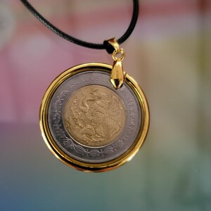 May include: A pendant necklace featuring a Mexican coin encased in a polished gold-tone circular frame. The coin displays a central gold-colored eagle emblem surrounded by a silver-colored border with the text ESTADOS UNIDOS MEXICANOS.