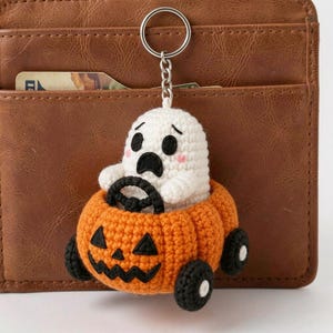 May include: A crochet keychain featuring a white ghost with black eyes and a surprised expression, sitting inside an orange pumpkin-shaped vehicle with black wheels and a black jack-o'-lantern face, attached to a silver metal keyring.